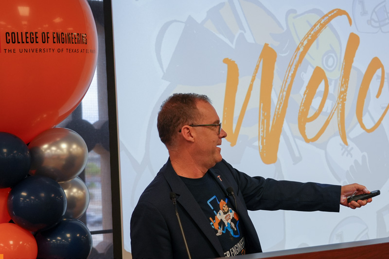 Two attendees wearing UTEP apparel and name badges pose together during a UTEP Engineering homecoming event, making hand gesture signs while standing in a banquet-style gathering space with other attendees visible in the background.